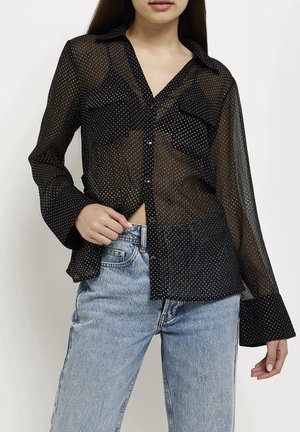 Schwarzes transparentes Hemd mit weißen Punkten, mit Kragen, Brusttaschen und langen Ärmeln mit weitem Bund. Über einer blauen Jeans getragen.