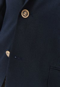 Granatowa marynarka z teksturowanej tkaniny, ozdobiona złotymi guzikami z wytłoczonym wzorem oraz jedną kieszenią z przodu.