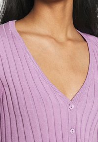 Närbild av en person som bär en ribbad lavendelfärgad V-ringad topp med två knappar synliga på bröstområdet.