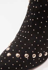 Mustat mokkanahkaiset nilkkurit, joissa on erikokoisia metallisia niittejä, terävä kärki ja sileä pinta, joka luo tyylikkään muotoilun.