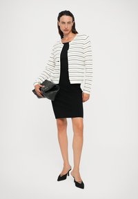 Bijela prugasta kardigan s otvorenim prednjim dijelom preko uske crne haljine, uparen s crnim štiklama i velikom crnom torbom. Elegantni, moderni dizajn.