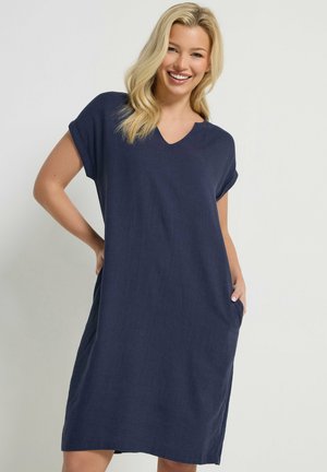 Uśmiechnięta kobieta o blond włosach, ubrana w luźną granatową sukienkę z dekoltem w serek, krótkimi rękawami, z rękami w kieszeniach, na jednolitym tle.