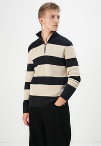Gestreifter Strickpullover in Marineblau und Beige, mit einem Viertelreißverschluss und geripptem Textur. Lange Ärmel und lässige Passform, kombiniert mit schwarzen Hosen.