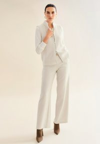 Frau in einem cremefarbenen, gestrickten, ärmellosen Oberteil über einem Langarmoberteil, cremefarbenen, weiten Hosen und braunen, spitzen Stiefeletten, die vor einem hellen Hintergrund steht.