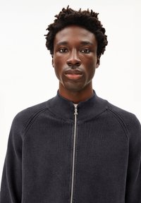 Donker marineblauwe gebreide jas met een hoge kraag, volledige rits en geribbelde textuur. Kenmerkt een naadloos raglanontwerp voor een getailleerde pasvorm.