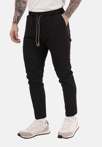 Pantalones de chándal negros con cintura elástica y cordón ajustable. Presentan piernas ajustadas y bolsillos laterales. Combinados con zapatillas blancas.