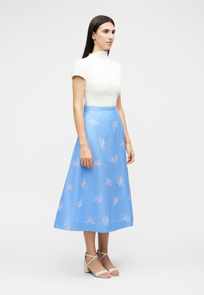 Γυναίκα όρθια, φοράει λευκό μπλουζάκι με κοντά μανίκια και ριμπ ύφανση, μπλε φούστα σε σχήμα Α με φλοράλ σχέδια και μπεζ σανδάλια με τακούνι, μπροστά από απλό φόντο.