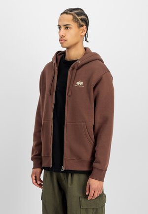 Junger Mann mit geflochtenem Haar, der einen braunen Kapuzenpullover mit Reißverschluss und grüne Cargohosen trägt, steht vor einem einfarbigen hellen Hintergrund.