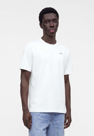 Giovane uomo con capelli corti e ricci, che indossa una semplice t-shirt bianca a maniche corte e jeans azzurri chiari, in piedi davanti a uno sfondo chiaro.