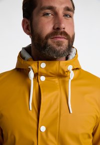 Gul vandtæt regnjakke med grå inderside, udstyret med hvide toggle-snore og knapdetaljer. Glat, blank tekstur.