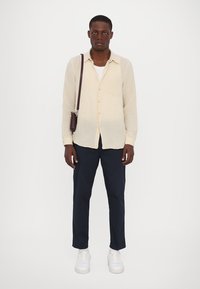 Camicia bottonata beige chiaro, pantaloni blu navy, sneakers bianche e una piccola borsa scura, indossata da un modello. La camicia ha una vestibilità comoda e un dettaglio della tasca.