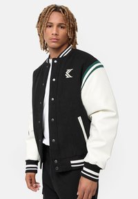Jachetă de tip varsity neagră și crem cu dungi verzi, nasturi tip snap, manșete ribbate și un logo brodat pe piept.