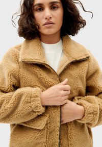 Fleece zip-up jack in lichtbruin met een gestructureerd, fluffy oppervlak. Voorzien van een hoge kraag, zakken aan de voorkant en een metalen ritssluiting.