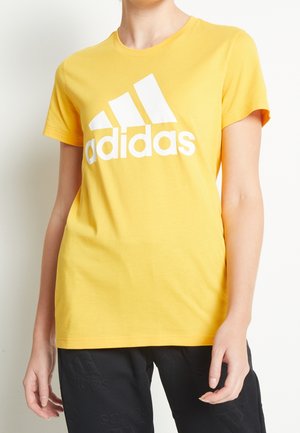 Person iført en gul Adidas t-shirt med stort hvidt logo og sorte bukser mod en ensfarvet lys baggrund.
