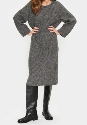 Kobieta ubrana w długą szarą dzianinową sukienkę z szerokimi rękawami i czarne wysokie skórzane kozaki, stojąca z rękami na biodrach.