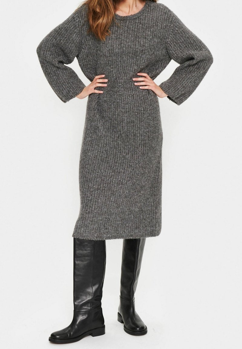 Femme portant une longue robe grise en maille avec des manches larges et des bottes en cuir noires montant jusqu'aux genoux, debout les mains sur les hanches.