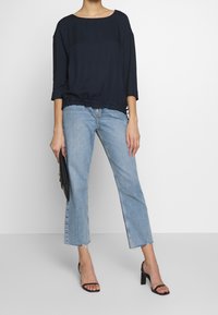 Frau, die ein lockeres navyblaues Oberteil, hellblaue cropped Jeans mit rohem Saum, schwarze High-heel-Sandalen trägt und eine schwarze Clutch-Tasche hält.