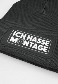 Black beanie with textured knit design. Features a rectangular patch with white text and a cat graphic, reading "ICH HASSE MONTAGE."