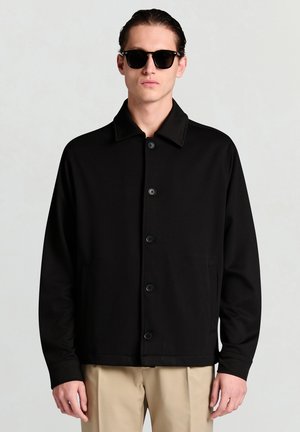 Schwarze Jacke mit Knopfleiste, einem strukturierten Kragen, zwei Seitentaschen und einer glatten Textur, kombiniert mit schwarzer Sonnenbrille und beigen Hosen.