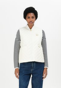 Gewatteerde witte vest met hoge kraag, gouden rits en accent, gedragen over een zwart-wit gestreepte langemouwen top, gecombineerd met spijkerbroek.