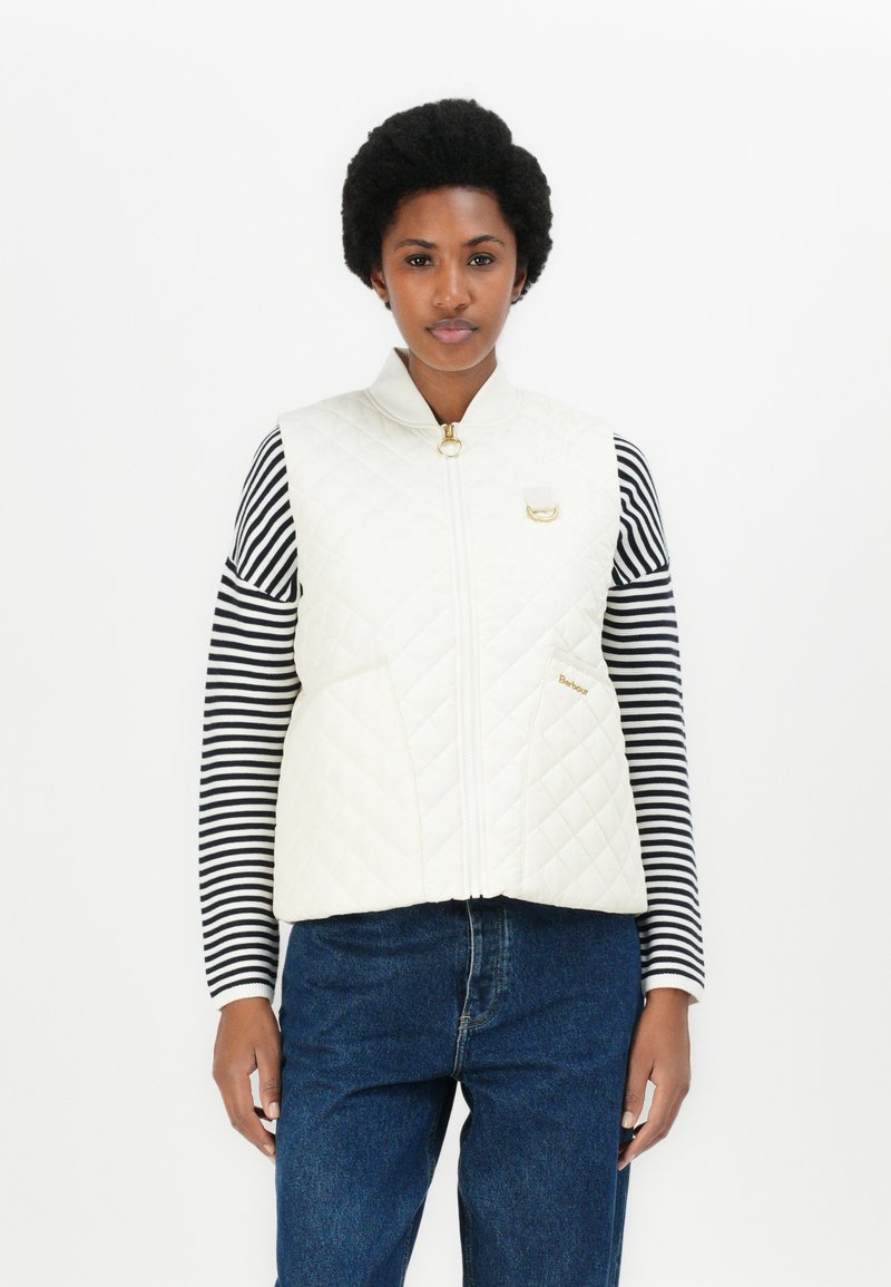 Gewatteerde witte vest met hoge kraag, gouden rits en accent, gedragen over een zwart-wit gestreepte langemouwen top, gecombineerd met spijkerbroek.