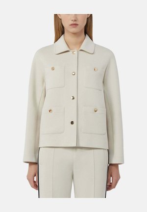 Vrouw draagt een crèmekleurig jasje met vier voorzakken en vierkante knopen in goudkleur, gecombineerd met bijpassende broek met zwarte zijstrepen.