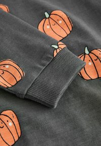 Grått tyg med ett upprepande mönster av orange pumpor med leende ansikten och gröna stjälkar. Ribbad mudd vid kanten. Mjuk textur.