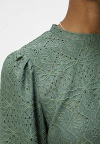 Grön broderad topp med texturerade blommönster, puffiga axlar och hög halsringning. Tyget har dekorativa utskärningar.