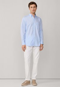 Camisa de botones azul claro de mangas largas, combinada con pantalones blancos y zapatos marrón claro. Presenta un bolsillo en el pecho y un cuello estándar.