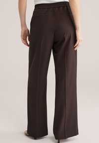 Vrouw die donkerbruine, wijde broek met hoge taille draagt, voorzien van een elastische tailleband en twee achterzakken, staand tegen een effen achtergrond.