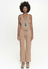 Beżowy kombinezon bez rękawów o luźnym kroju i regulowanym pasie; uzupełniony długim naszyjnikiem z koralików z niebieskim frędzlem.