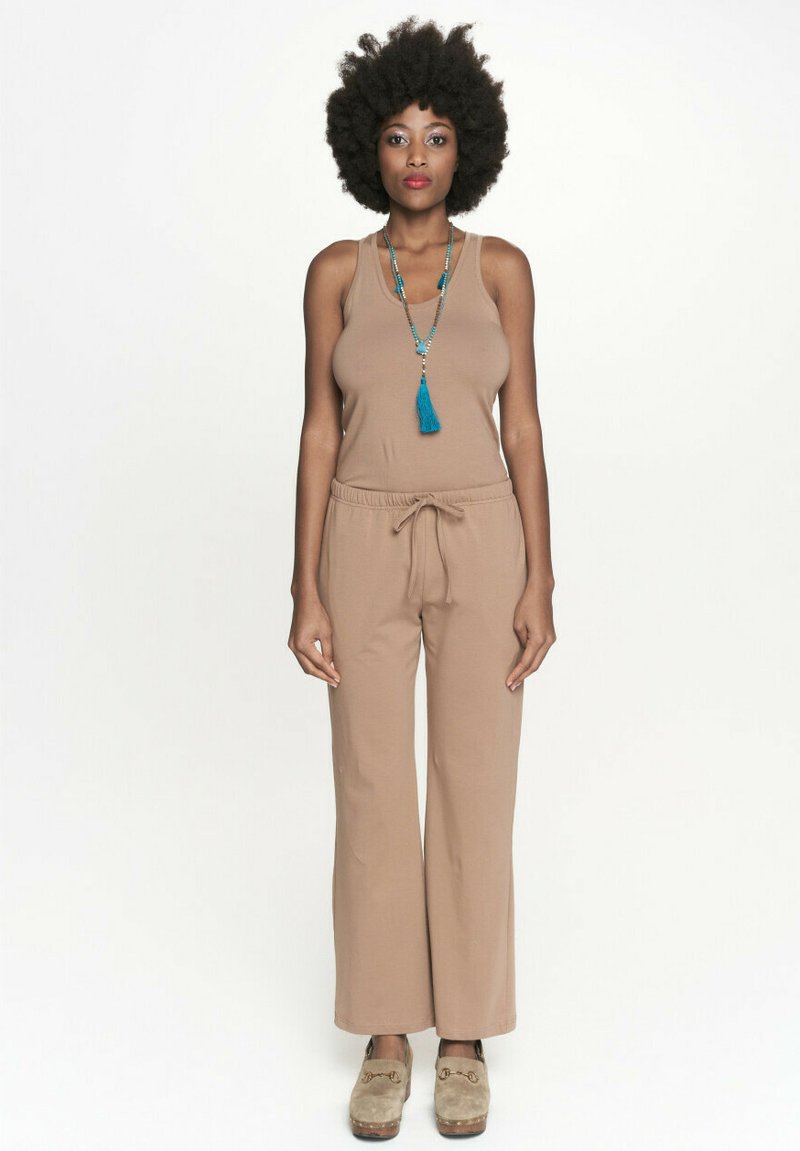 Beżowy kombinezon bez rękawów o luźnym kroju i regulowanym pasie; uzupełniony długim naszyjnikiem z koralików z niebieskim frędzlem.