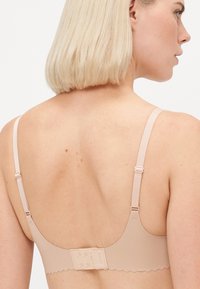Light beige bra with adjustable straps, scalloped edges, and a hook-and-eye closure at the back, made from smooth, stretchy material.