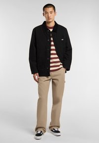 Černá plátěná bunda s knoflíkovým zapínáním, dvě přední kapsy, přes maroonovou a krémovou pruhovanou tričko, spárovaná s khaki kalhotami a černými teniskami.