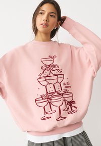 Ružová oversize mikina s veselou grafikou navrstvených pohárov a stúh v bordovej farbe. Má okrúhly výstrih a rebrované manžety.