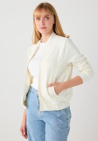 Crèmekleurig bomberjack met een gestructureerd ontwerp, ritssluiting en zijzakken. Gecombineerd met een wit T-shirt en lichtblauwe jeans.