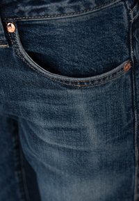Gros plan sur une poche avant de jean en denim bleu foncé avec des rivets en cuivre et des détails de couture visibles.