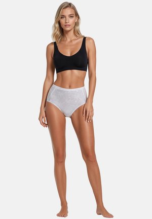 Frau steht barfuß, trägt einen schwarzen Sport-BH und hoch taillierte weiße Spitzenunterwäsche mit floralem Muster vor einem einfarbigen Hintergrund.