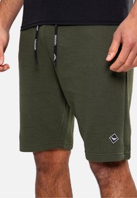 Olivgrüne Shorts mit strukturiertem Design, ausgestattet mit schwarzen Kordeln und einem kleinen weißen Logoetikett am unteren Saum.