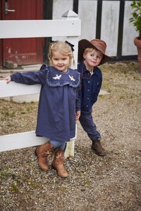 Tumši zila kleita ar rūtainu rakstu, baltu putna izšuvumu un garām piedurknēm. Brūni kovboju zābaki ar izsmalcinātu šuvumu. Bērns tur roku rokā.