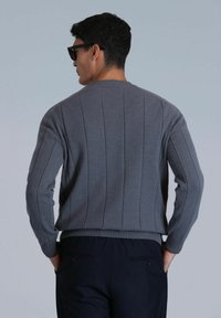 Pull en maille gris avec un design à côtes verticales, encolure ronde et manches longues, associé à un pantalon foncé. Tissu simple et texturé.