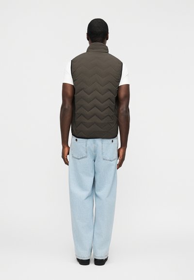 Άνδρας που φοράει σκούφο κοντό μπουφάν πάνω από λευκό μπλουζάκι και ανοιχτόχρωμο μπλε τζιν, στέκεται με την πλάτη προς τα εμπρός σε ένα απλό φωτεινό φόντο.