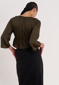 Maslinasto zelena satenasta bluzica s peplum rubom i rukavima do tri četvrtine, nosi se sa crnom traper suknjom visokog struka koja ima stražnje džepove.