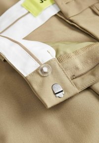 Pantalons beiges avec une taille blanche et une étiquette verte, dotés d'une pince en métal et d'un bouton transparent. Tissu doux avec une texture lisse.