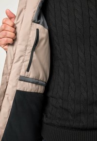 Una persona che indossa un maglione nero a trecce tiene aperta una giacca beige e nera, mostrando la tasca interna con zip della giacca.