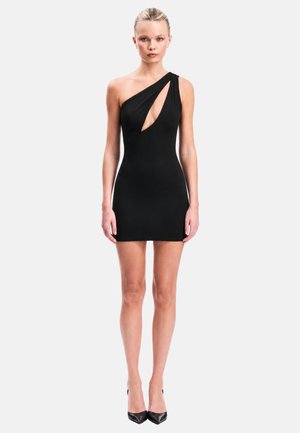 Blonde Frau trägt ein eng anliegendes schwarzes Minikleid mit einem diagonalen Ausschnitt an einer Schulter, steht mit entspannten Armen und trägt schwarze High Heels.
