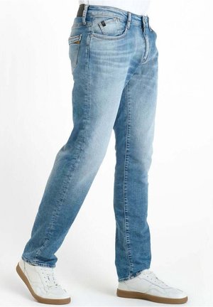 Hellblaue ausgewaschene Jeans, getragen mit weißen Sneakern und einem schwarzen Gürtel, von der Taille bis zu den Füßen vor einem einfarbigen weißen Hintergrund gezeigt.