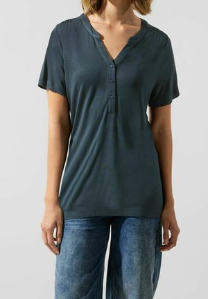 Femme portant une chemise bleu foncé à manches courtes boutonnée et un jean bleu, debout devant un fond clair uni.