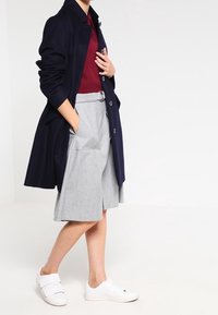 Marinblå lång kappa över en vinröd topp, parat med grå skräddarsydda shorts och vita sneakers. Kappan har krage och knappar.