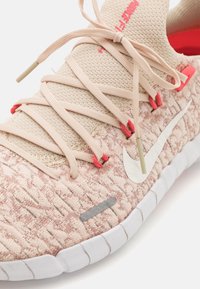 Lichtroze mesh sneaker met een gestructureerde oppervlakte, beige veters, rode accenten en een wit Nike-logo; heeft een flexibele zool.